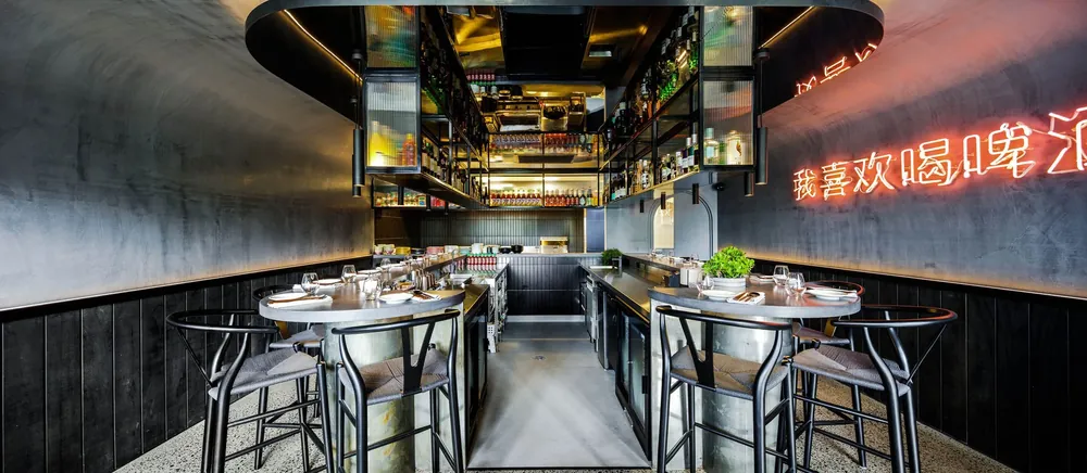 Modern bar with black walls, neon sign in Asian characters, metal tables with glasses and plates, and an overhead shelf stocked with liquor bottles.