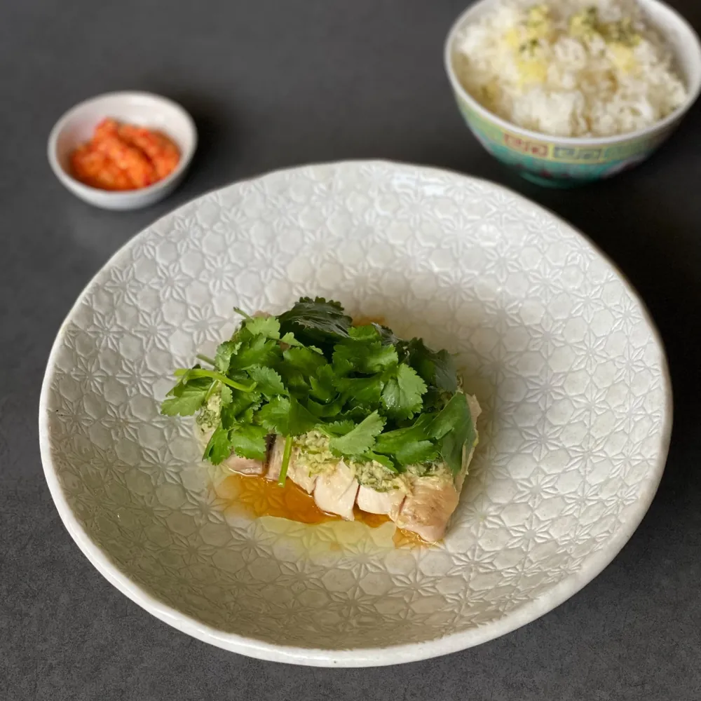 A dish with raw fish topped with herbs and sauce, served in a white bowl with a textured pattern. In the background, there are small bowls of condiment and a larger bowl of rice.