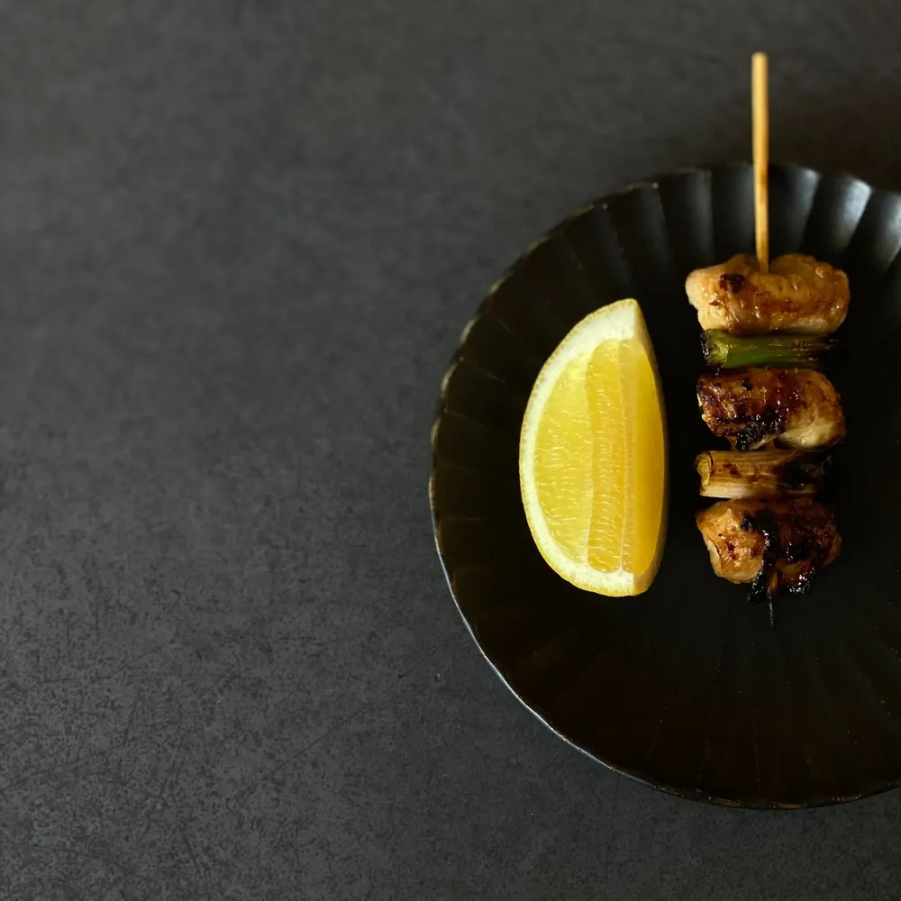 A black plate with two pieces of grilled meat on a skewer and a lemon wedge on a dark surface.